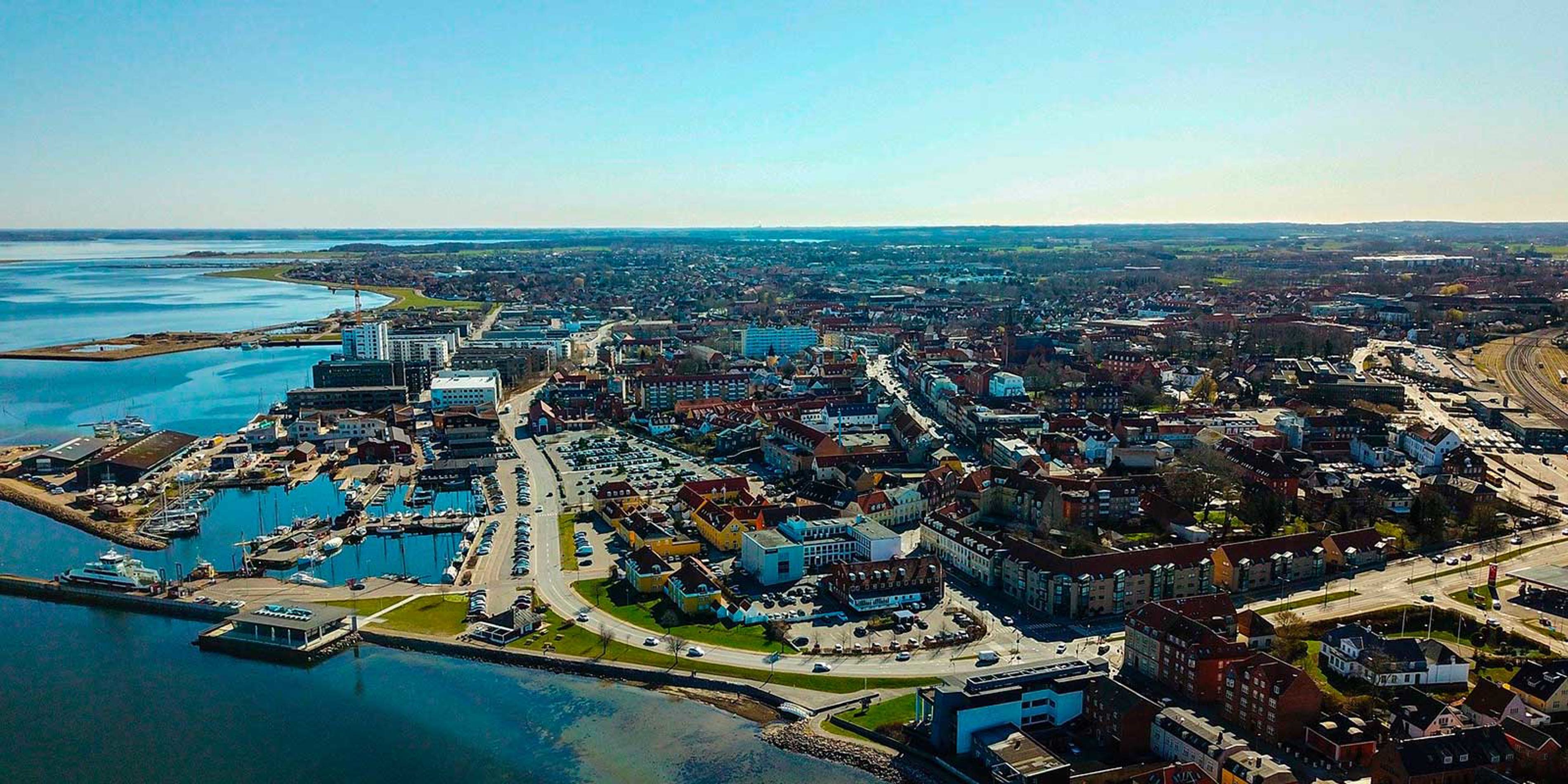 Flyfotografi af bybilledet i Horsens med havneområde og bygninger ved kysten på en solrig dag.