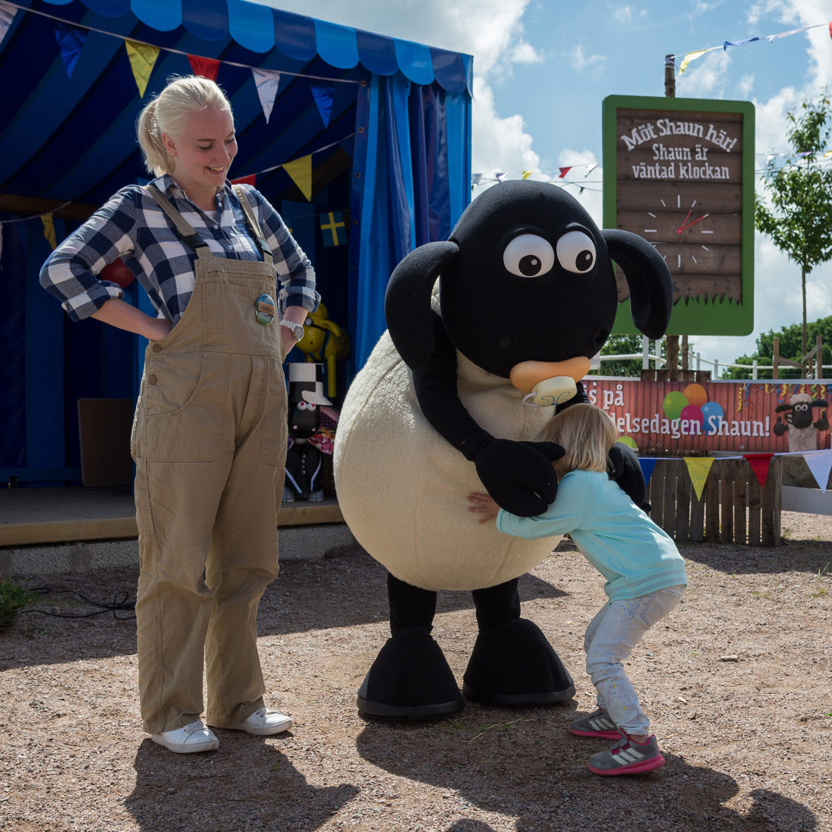 Barn omfavner en stor, sort og hvid fåremaskot ved et udendørs arrangement under en solrig himmel.