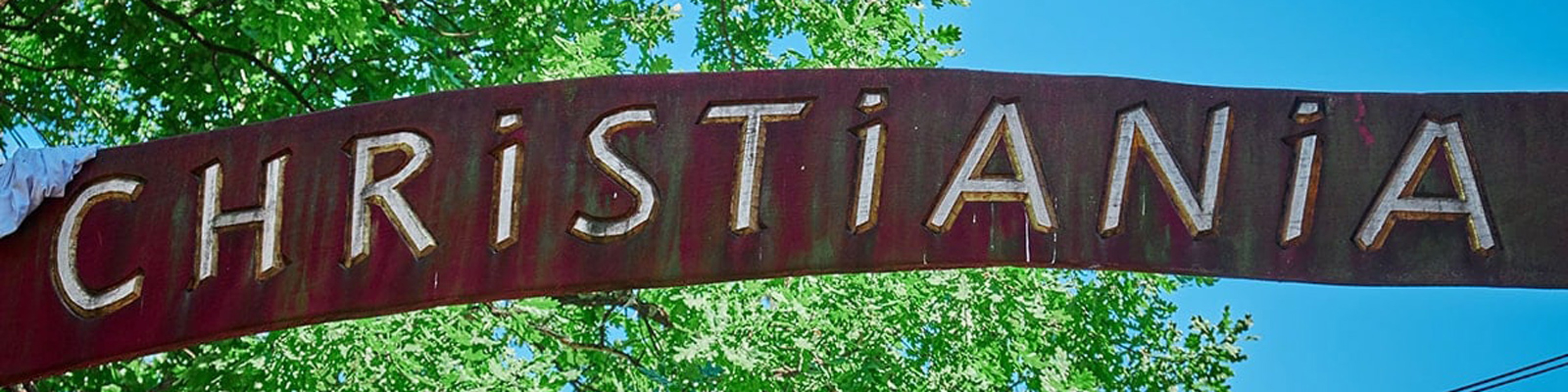 Skylt med texten "Christiania" i trä mot en bakgrund av gröna träd och blå himmel.