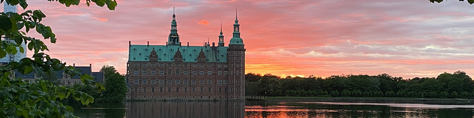 Frederiksborgs slott sett framifrån vid solnedgång med röd himmel och lugnt vatten i förgrunden.