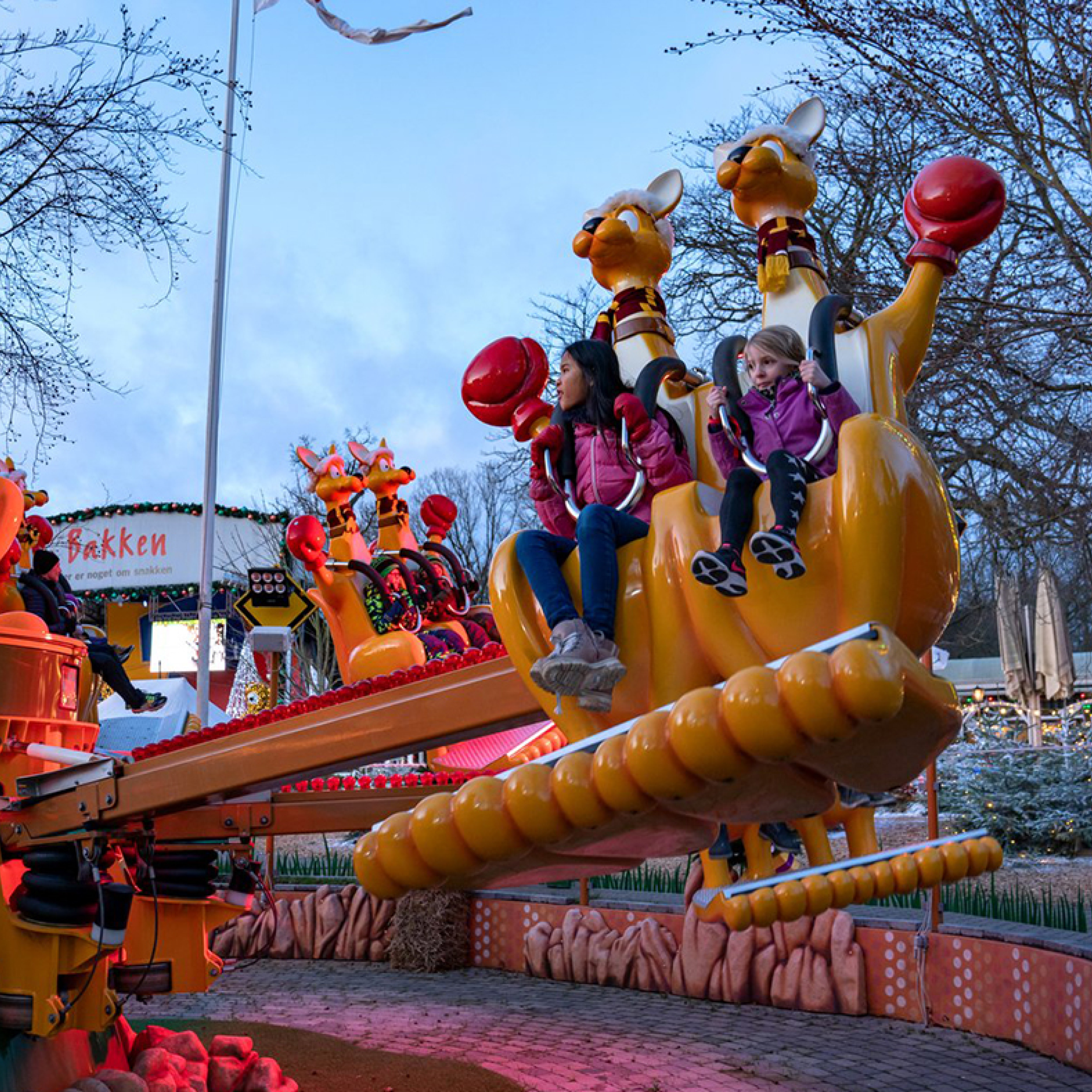 To piger i pink jakker på en gul forlystelse i Bakken forlystelsespark, omgivet af farverige kænguruer.
