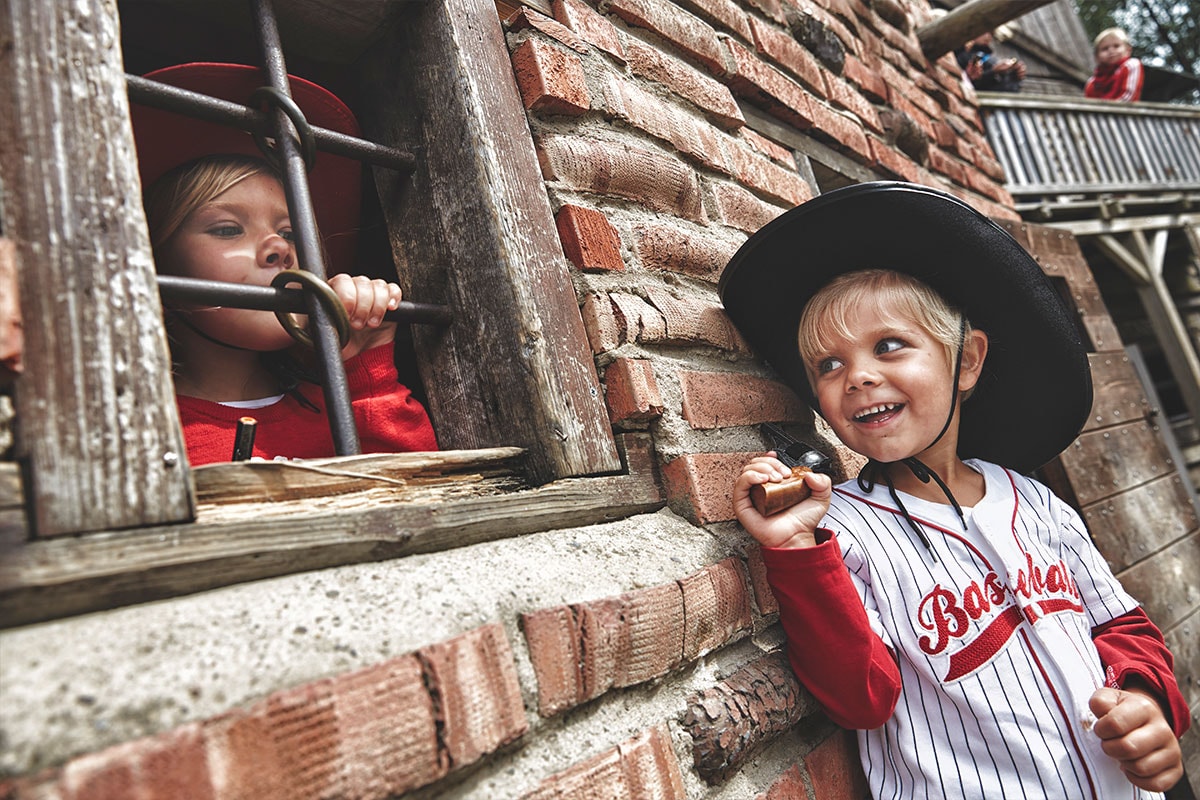 To børn står på hver deres side af et vindue med tremmer, begge iført cowboyhat.