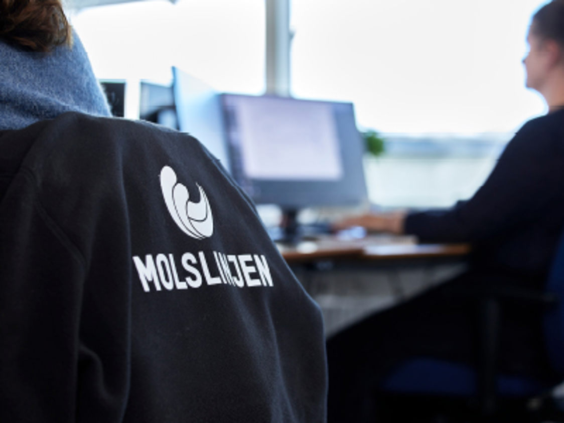 Medarbejder iført Molslinjen jakke arbejder ved et skrivebord med computerskærm i kontormiljø.