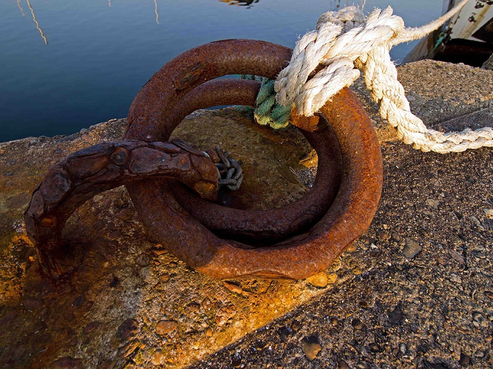 Rostig järnring med rep mot hamnens kajkant i närbild.