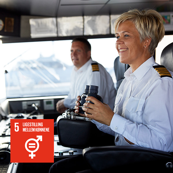 Kvinde kaptajn i hvid uniform styrer skib på broen med mandlig kollega i baggrund, fokus på ligestilling.