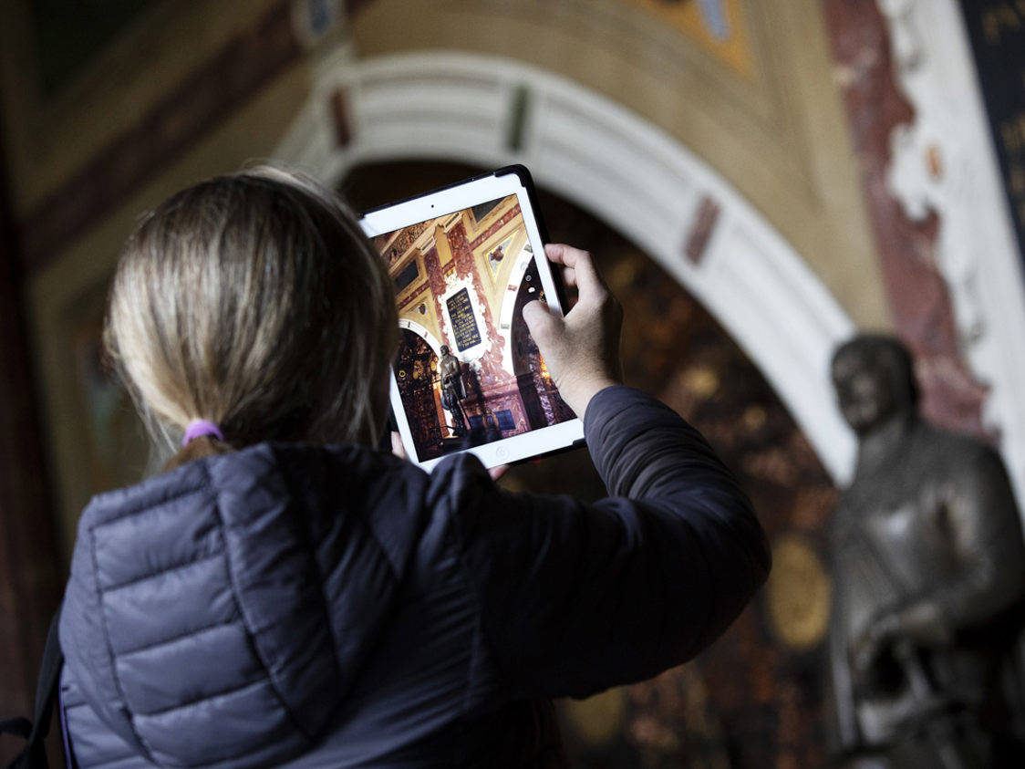 En person tager et foto af et historisk kirkerum med en tablet, set bagfra.