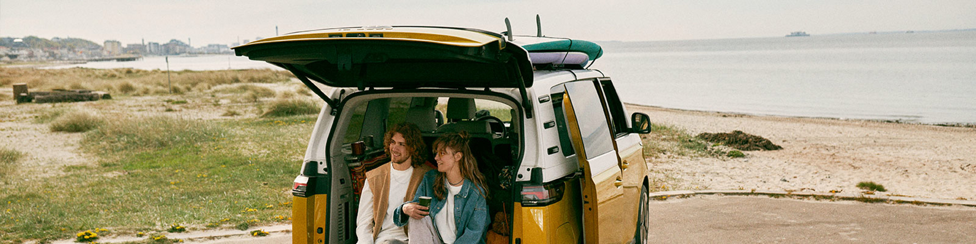 Yellow Surf Minibus on a coastal road with surfboards on roof, parked near the beach under a clear sky.