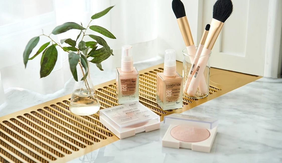 Makeup-sæt med foundation og børster på marmorplade ved siden af dekorativ vase med blade.