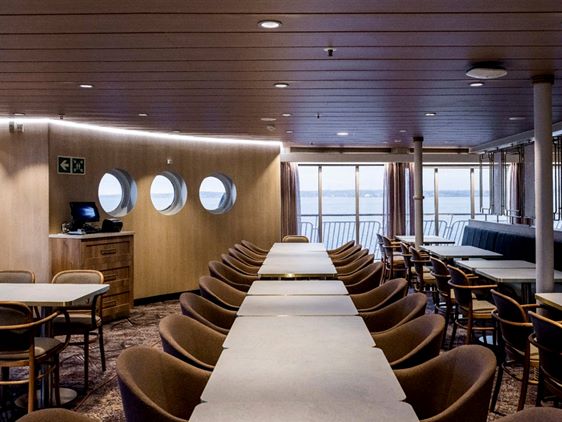 Færgens spiseområde med runde vinduer, træpaneler og stole ved borde med havudsigt gennem vinduerne.