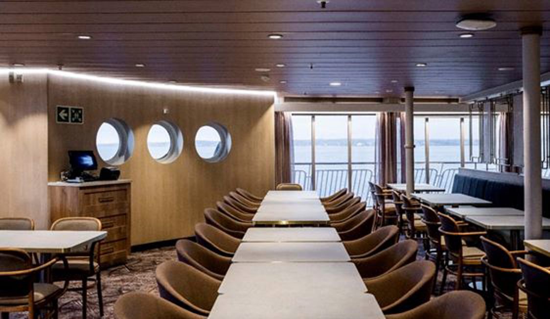 Færgens spiseområde med runde vinduer, træpaneler og stole ved borde med havudsigt gennem vinduerne.