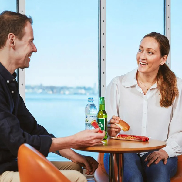 To mennesker nyder frokost ved et bord med sodavand på en Molslinjen færge med havudsigt gennem vinduet.