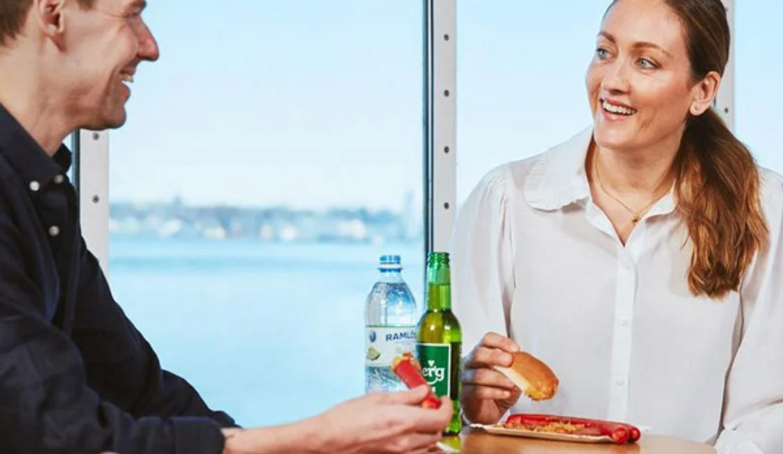 Två personer njuter av lunch med läsk vid ett bord på en Molslinjen-färja med utsikt över havet genom fönstret.