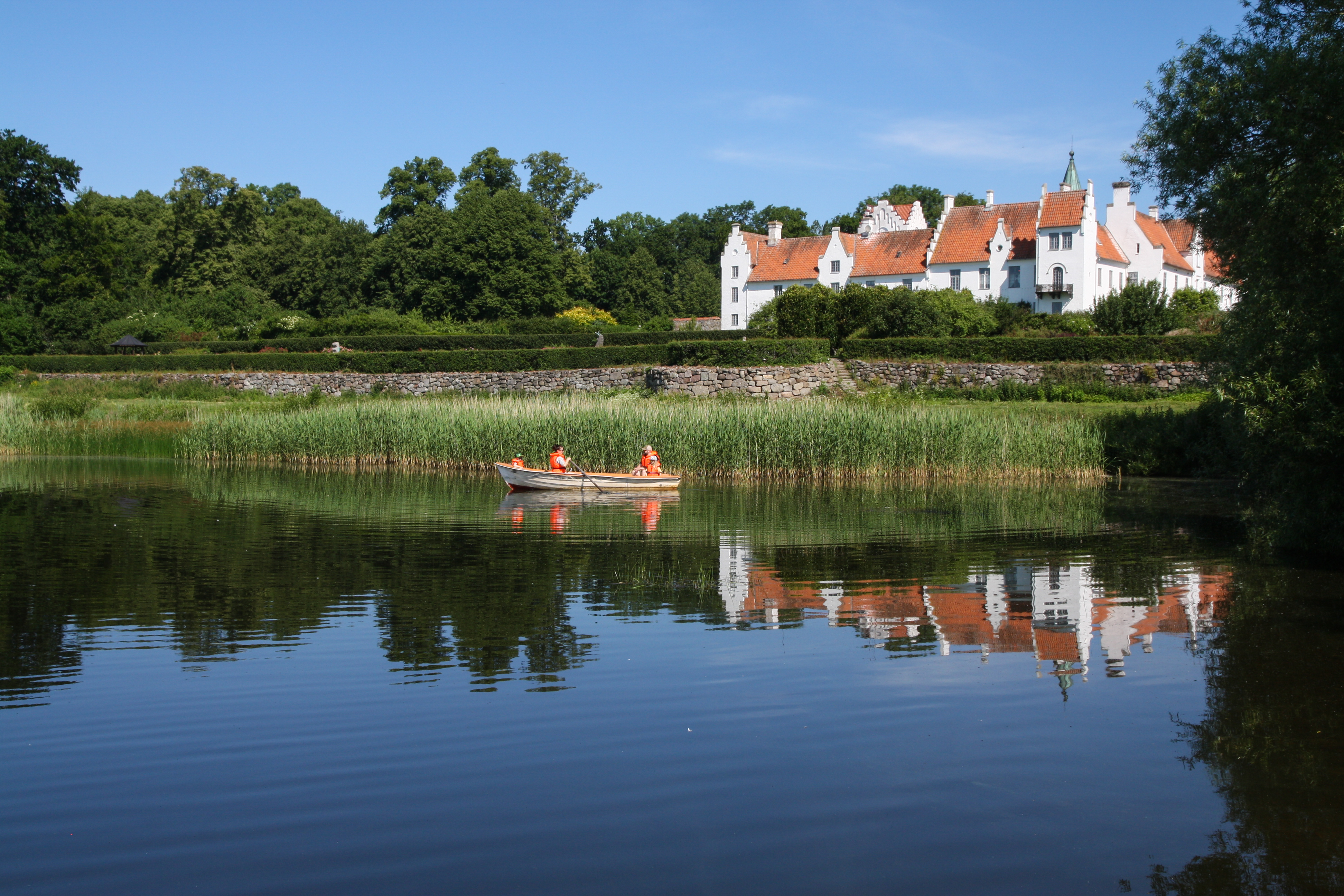 Robåd med personer i orange veste på en sø foran et historisk hvidt slot i solrigt vejr.