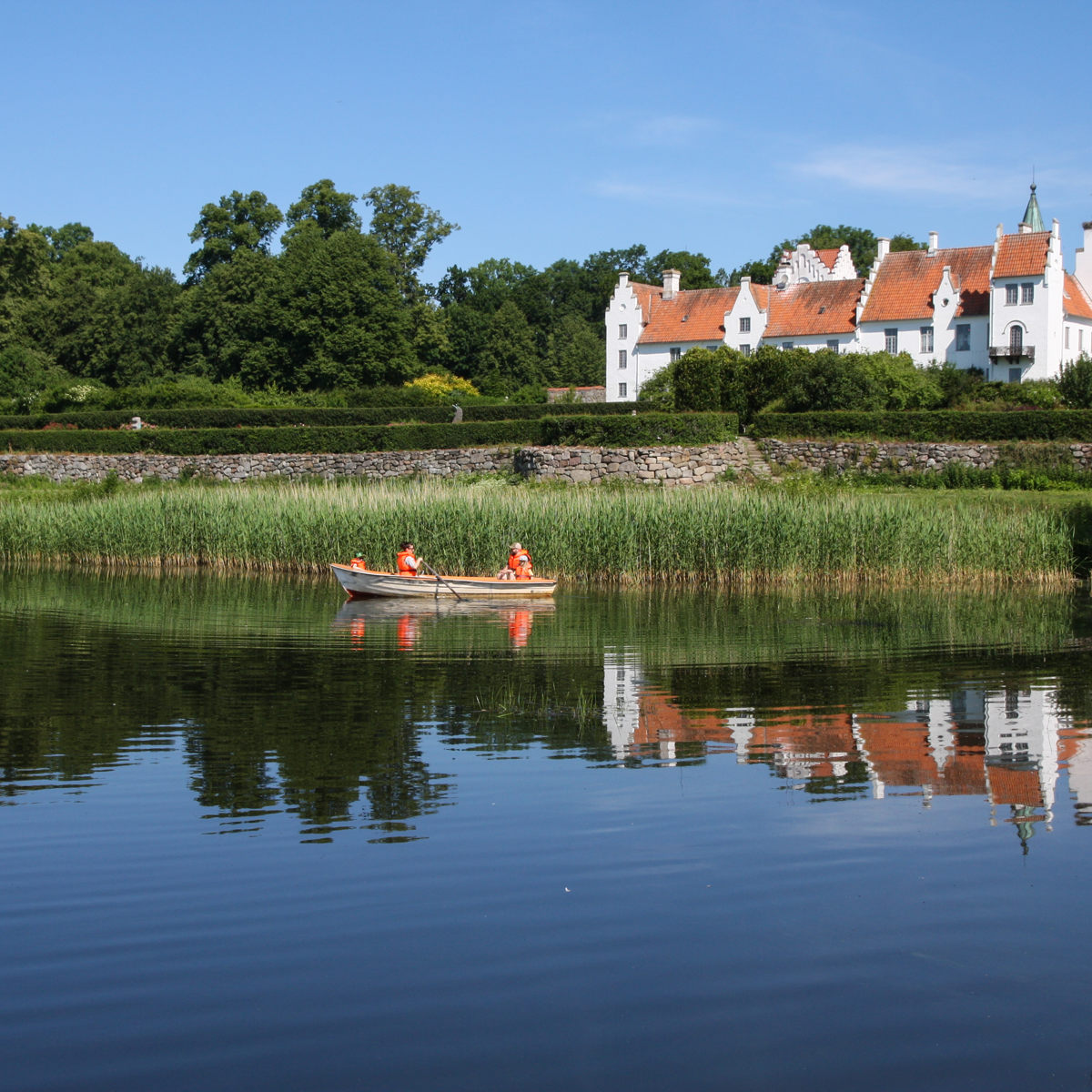 Robåd med personer i orange veste på en sø foran et historisk hvidt slot i solrigt vejr.