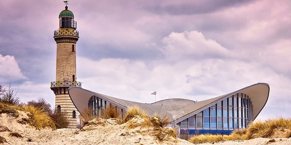 Warnemünde fyr vid stranden under en dramatisk rosa himmel med modern byggnad bredvid.