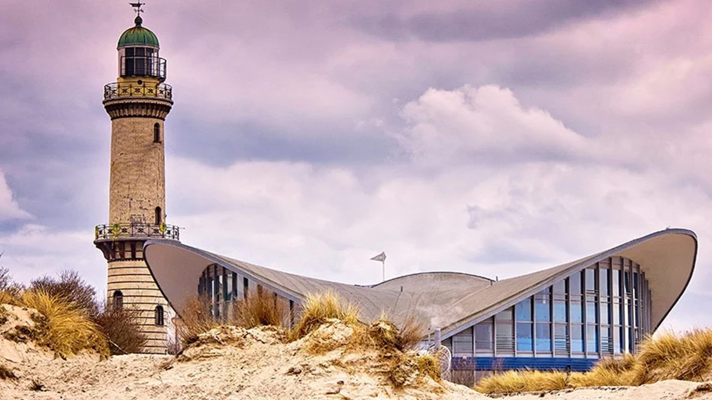 Warnemünde fyr vid stranden under en dramatisk rosa himmel med modern byggnad bredvid.