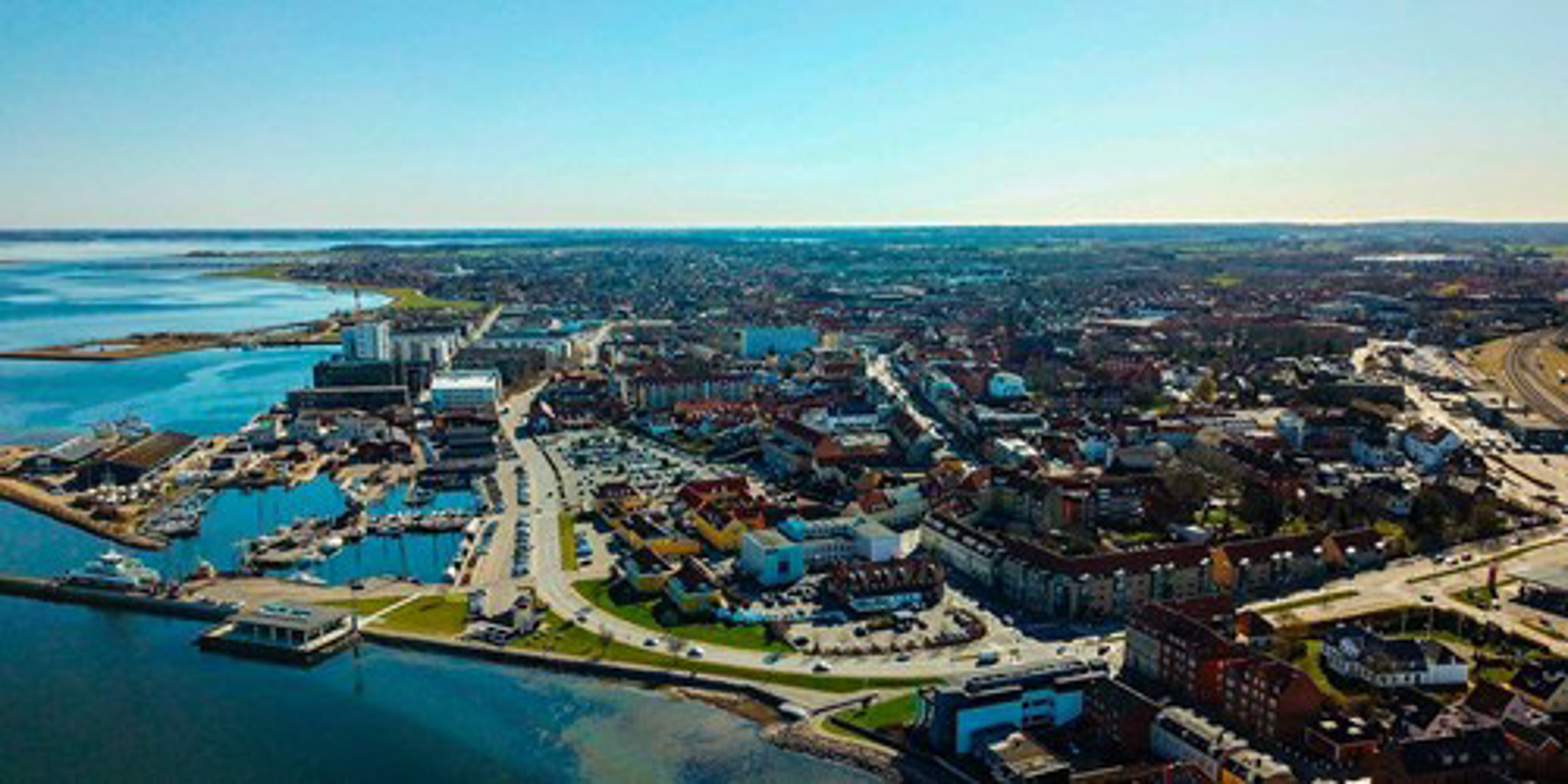 Luftfoto af bymidten i Esbjerg med udsigt over havnen og byens infrastruktur under en klar blå himmel.