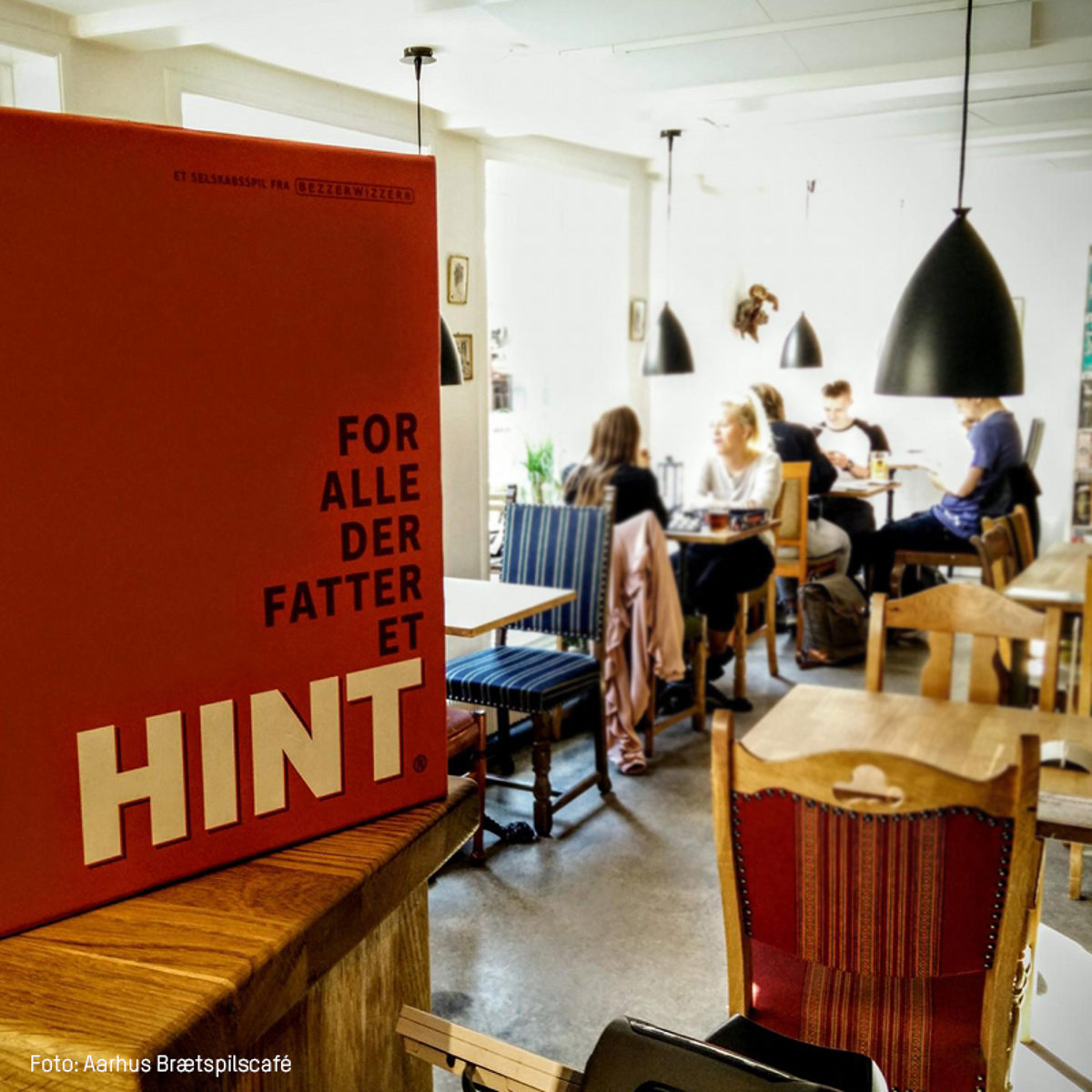 Brætspillet HINT vises i forgrunden på café, mens gæster sidder ved borde i baggrunden.