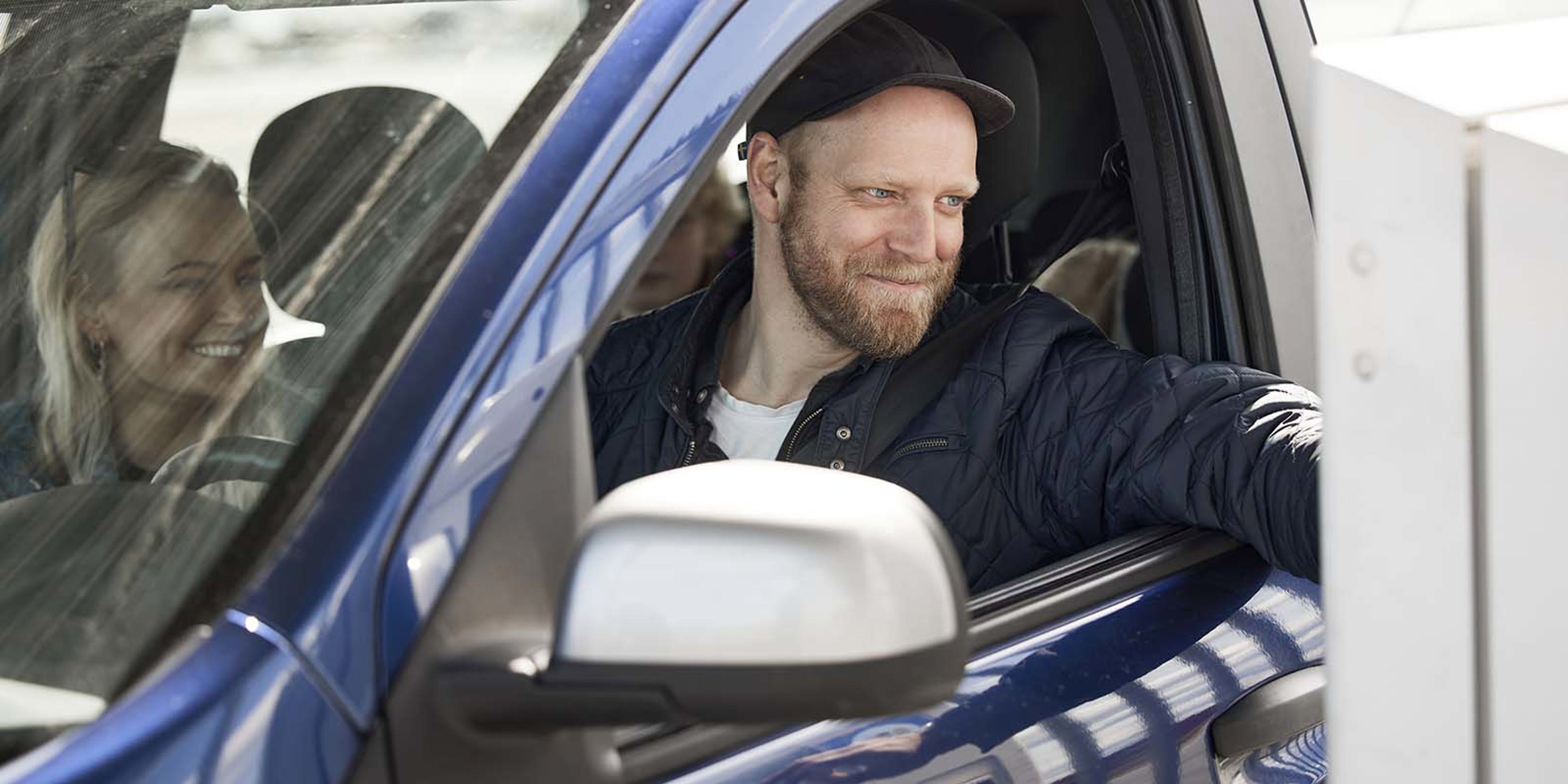Mand kører igennem betalingsanlæg i en blå bil og smiler, mens han betaler ved vinduet.