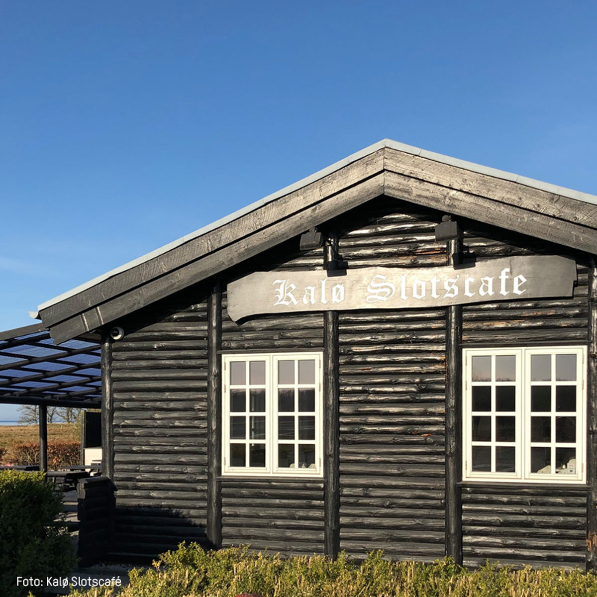 Kalø Slotscafe træhytte med mørkt træ og hvide vinduer under en klar blå himmel.