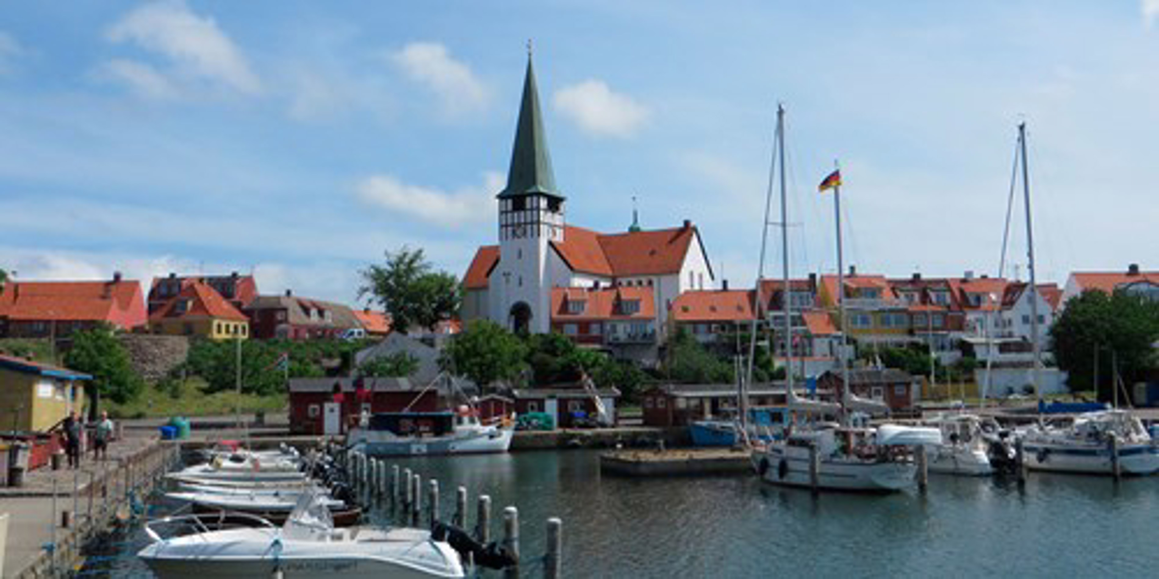 Kirke med spiret tårn og røde tage ved havn i solrig by, set fra vandet i fugleperspektiv.