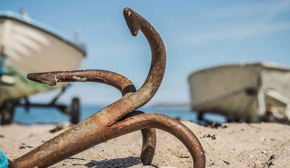 Rostigt ankare på sandstrand med suddiga båtar i bakgrunden i Liseleje, Nordsjälland.