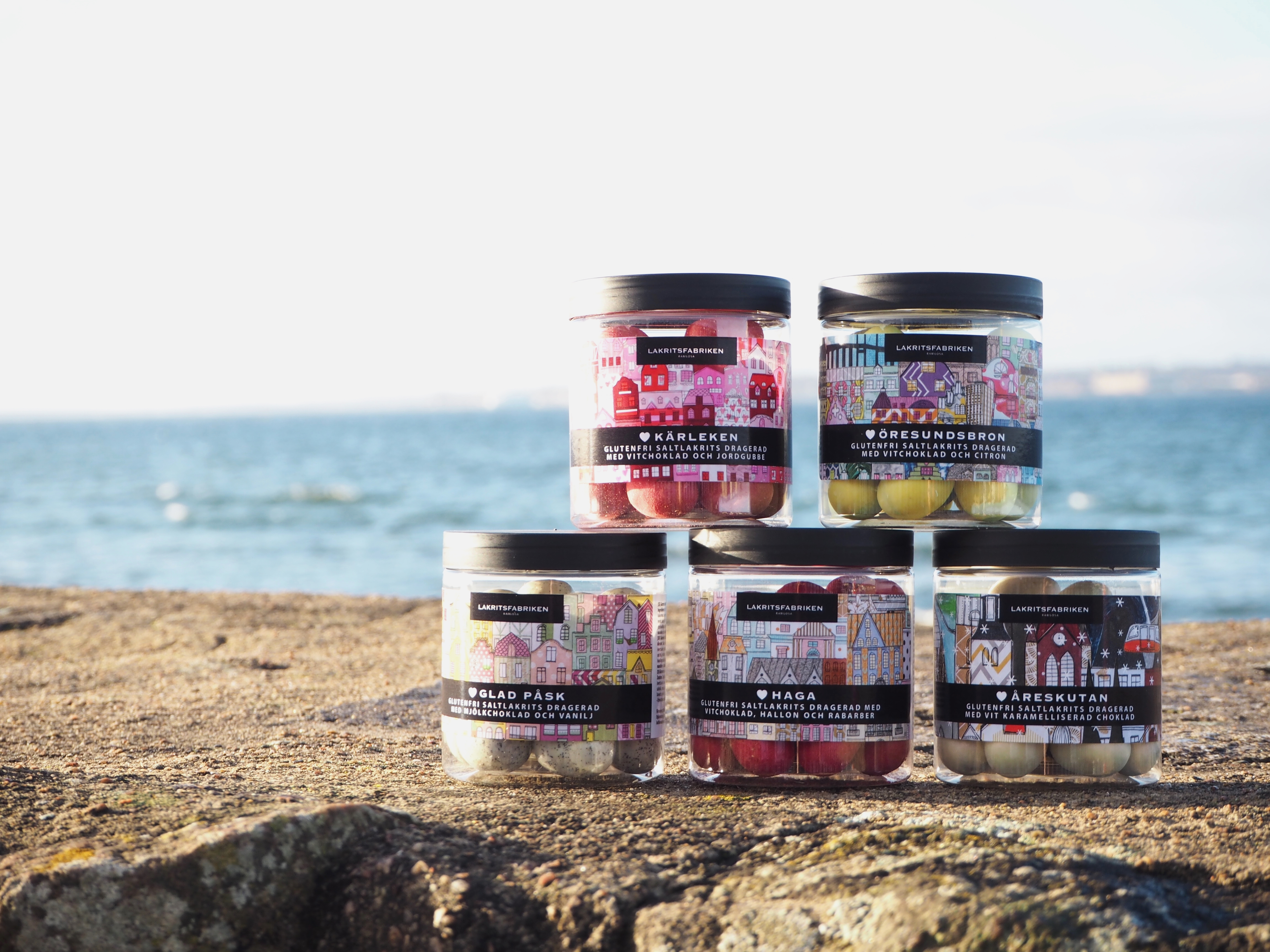 Fem krukker med lakrids placeret på en klippe med havudsigt i baggrunden, Lakritsfabriken mærkeartikler.