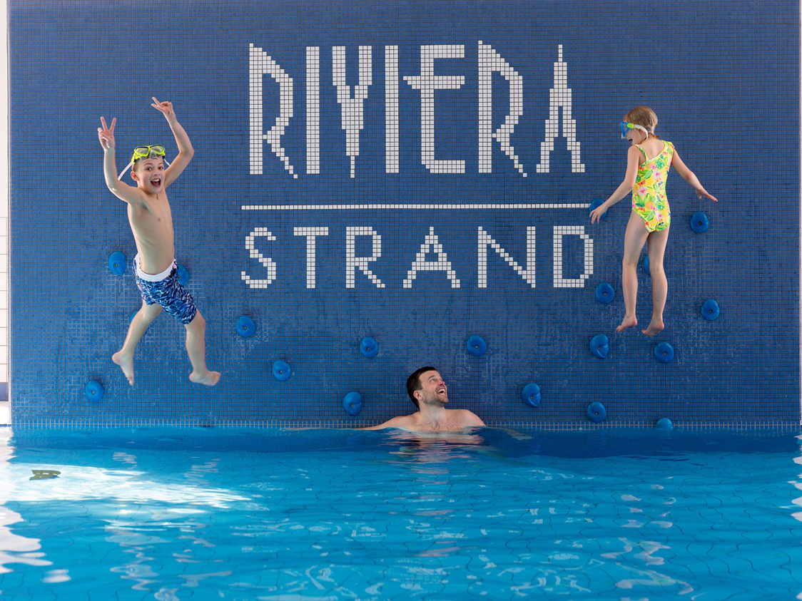 Børn leger i swimmingpool ved Riviera Strand, mens en voksen ser på, omgivet af blå fliser og klatregreb.