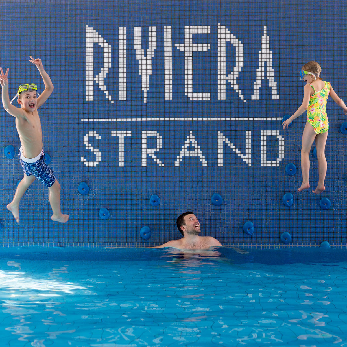 Børn leger i swimmingpool ved Riviera Strand, mens en voksen ser på, omgivet af blå fliser og klatregreb.