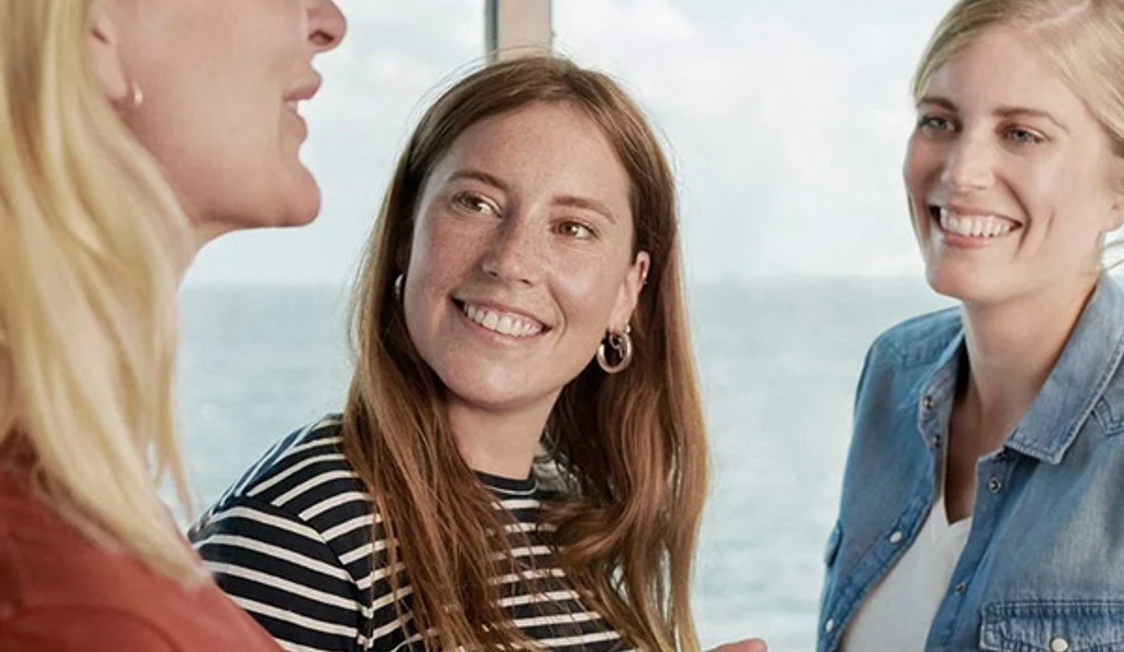 Tre kvinnor i samtal ombord på en färja med havsutsikt genom fönstret.