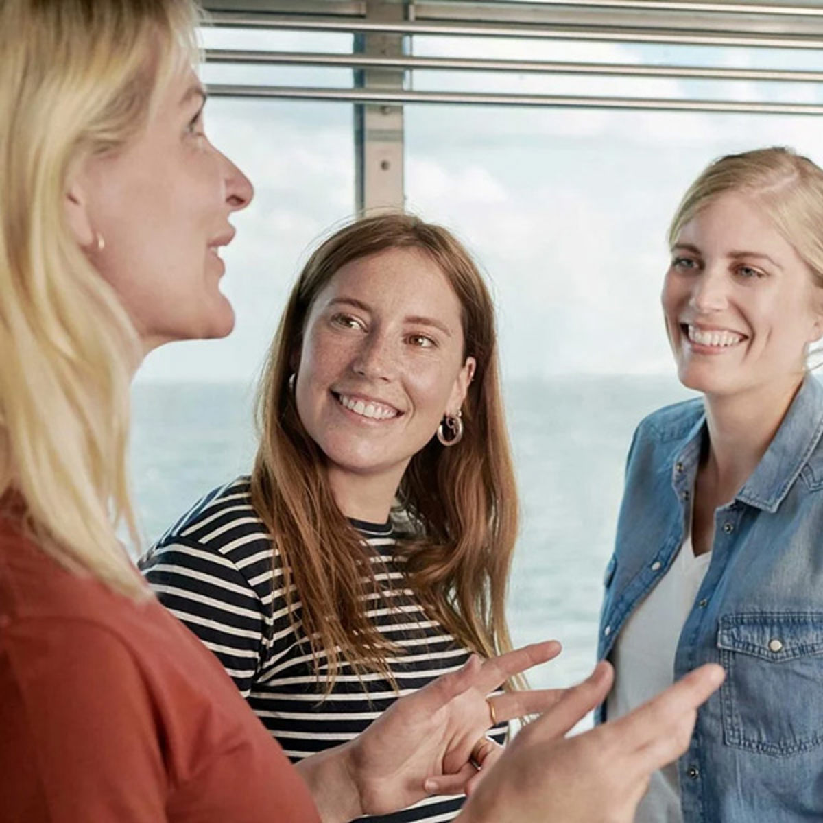 Tre kvinnor i samtal ombord på en färja med havsutsikt genom fönstret.