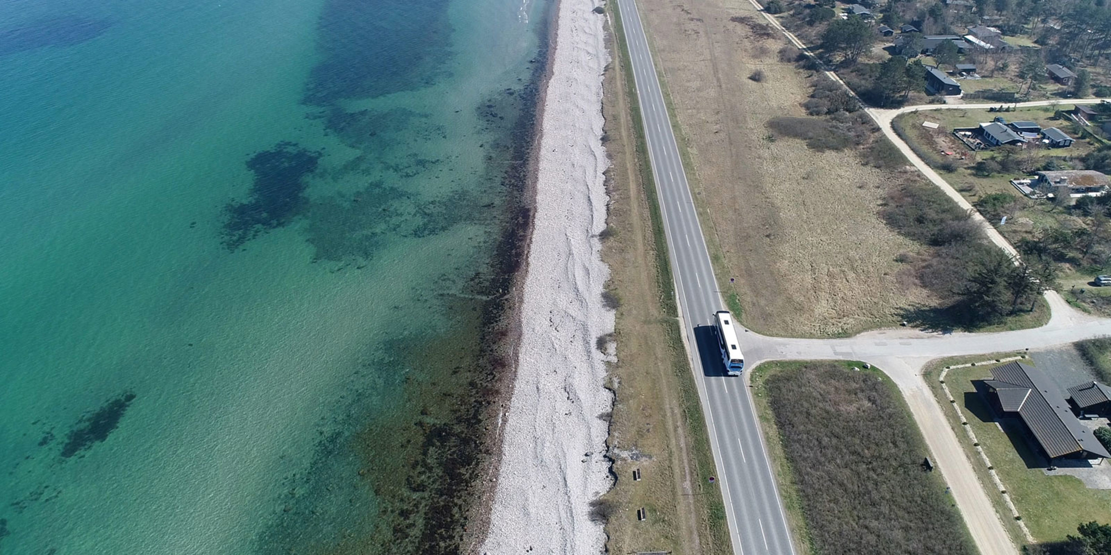 Kombardo Expressen bus travels along coastal road beside beach with clear water, seen from above.