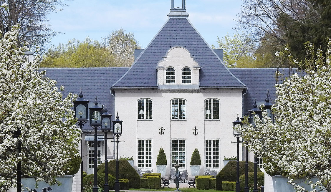 Historisk herregård med blomstrende træer og velplejet have i forgrunden på en solskinsdag.