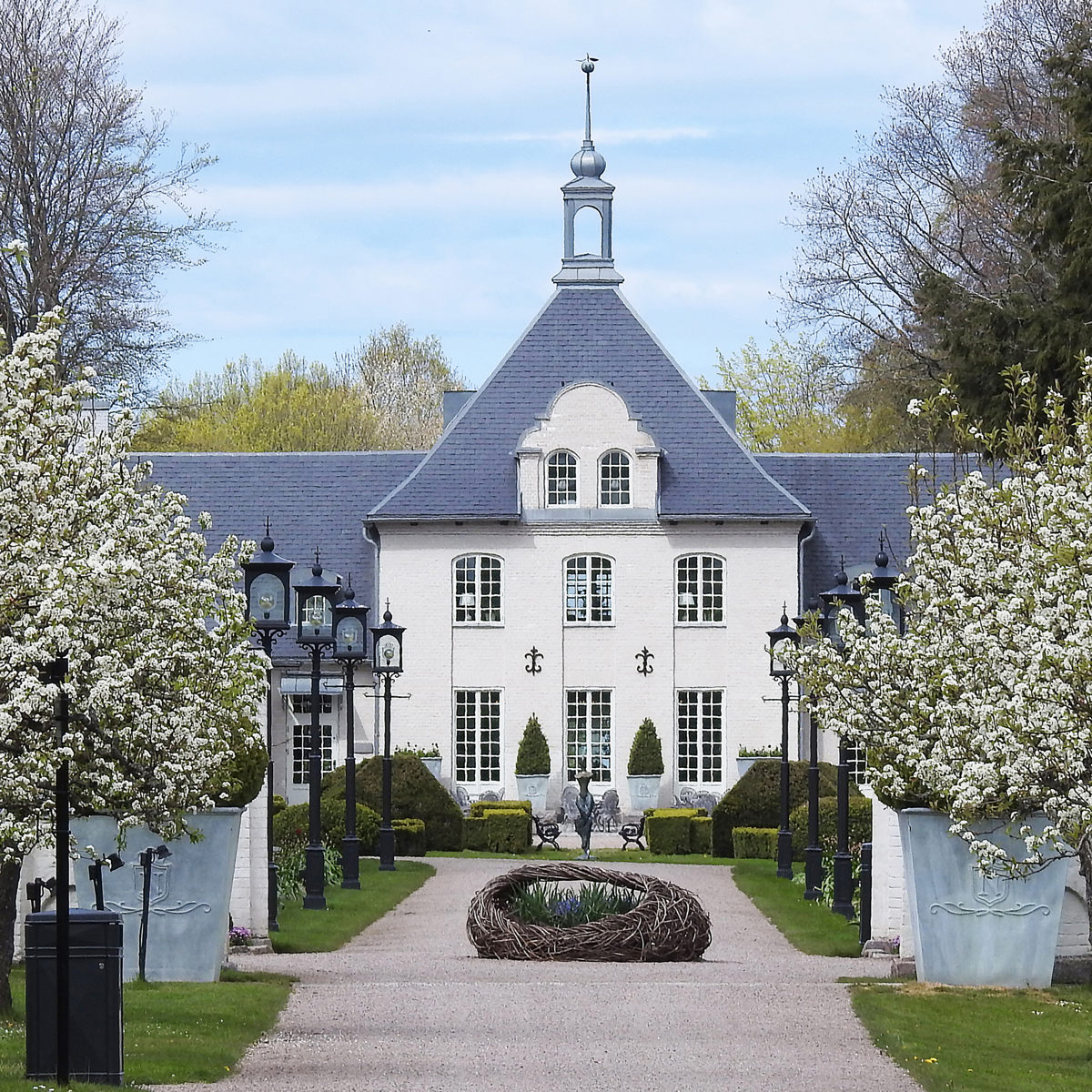 Historisk herregård med blomstrende træer og velplejet have i forgrunden på en solskinsdag.