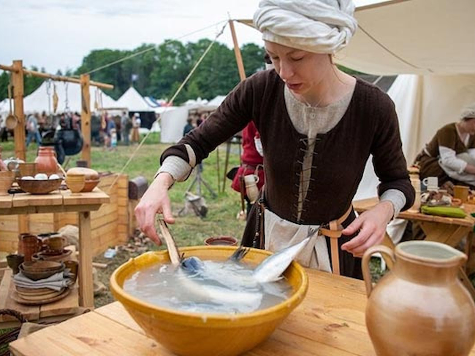 Kvinna klädd i medeltidsdräkt vid en marknadsplats hanterar fisk i en stor skål på ett träbord.