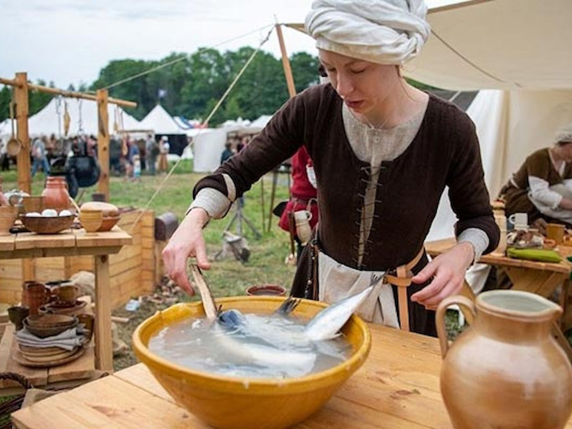 Kvinna klädd i medeltidsdräkt vid en marknadsplats hanterar fisk i en stor skål på ett träbord.