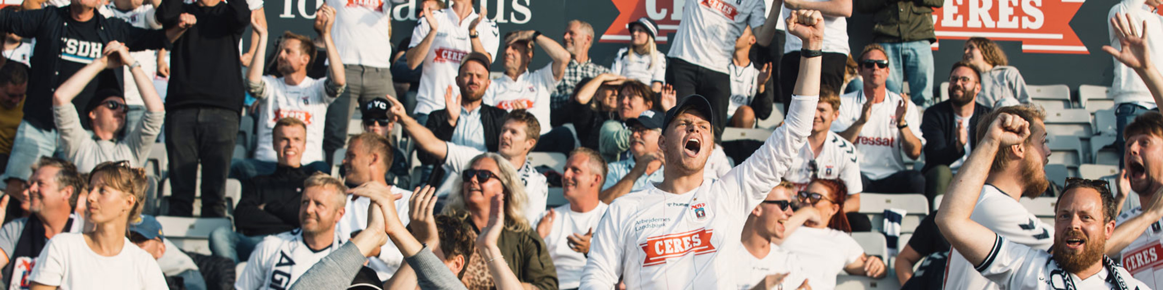 Fodboldfans i hvide trøjer jubler energisk på tribunerne under en spændende kampdag ved stadion.