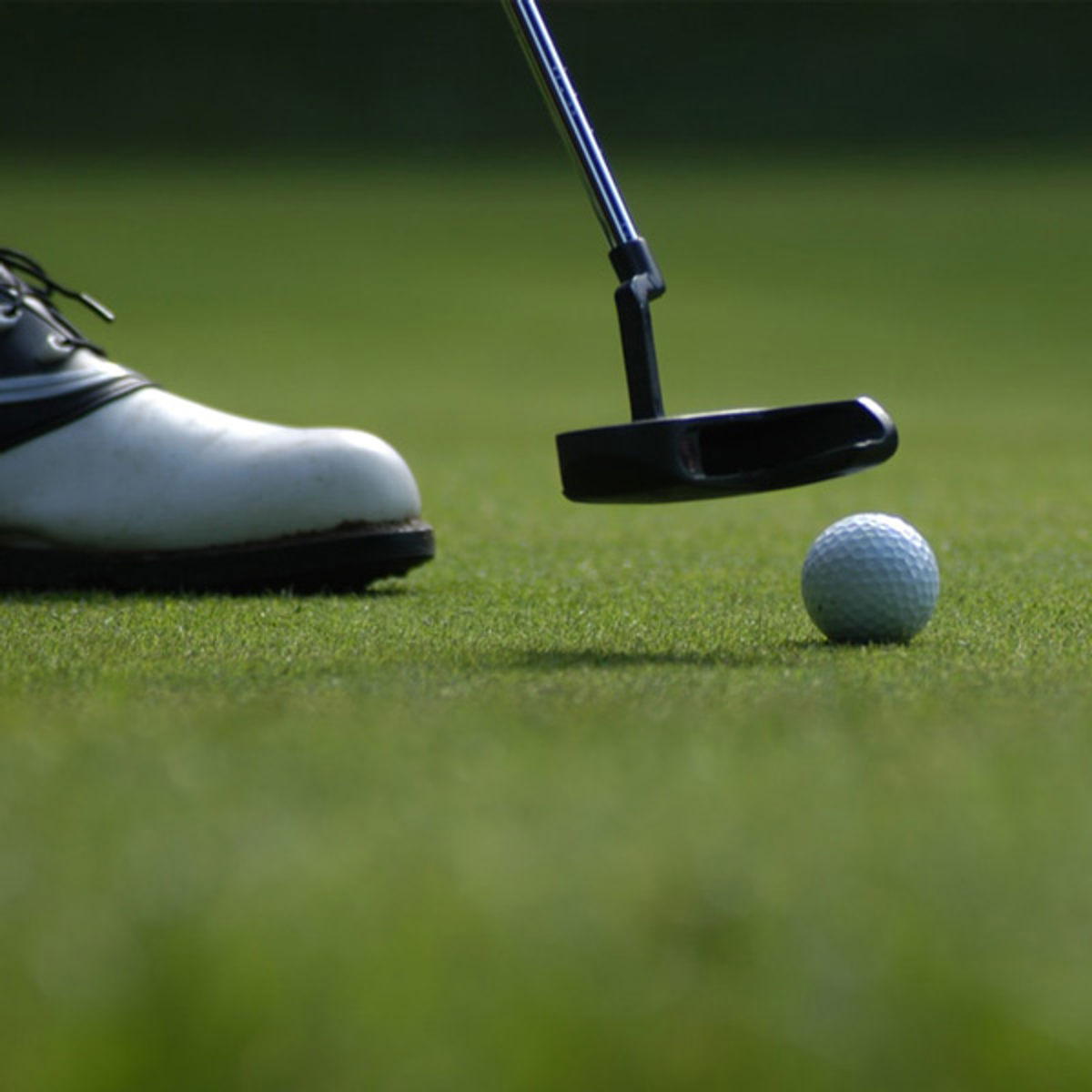 Golfspiller med hvid sko tager sigte mod golfbold med sort putter på grøn fairway i næroptagelse.