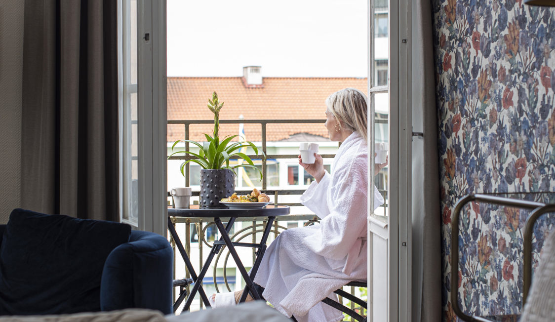 Kvinde iført hvid badekåbe nyder morgenkaffe og bagværk på balkon med udsigt til tag og plante.