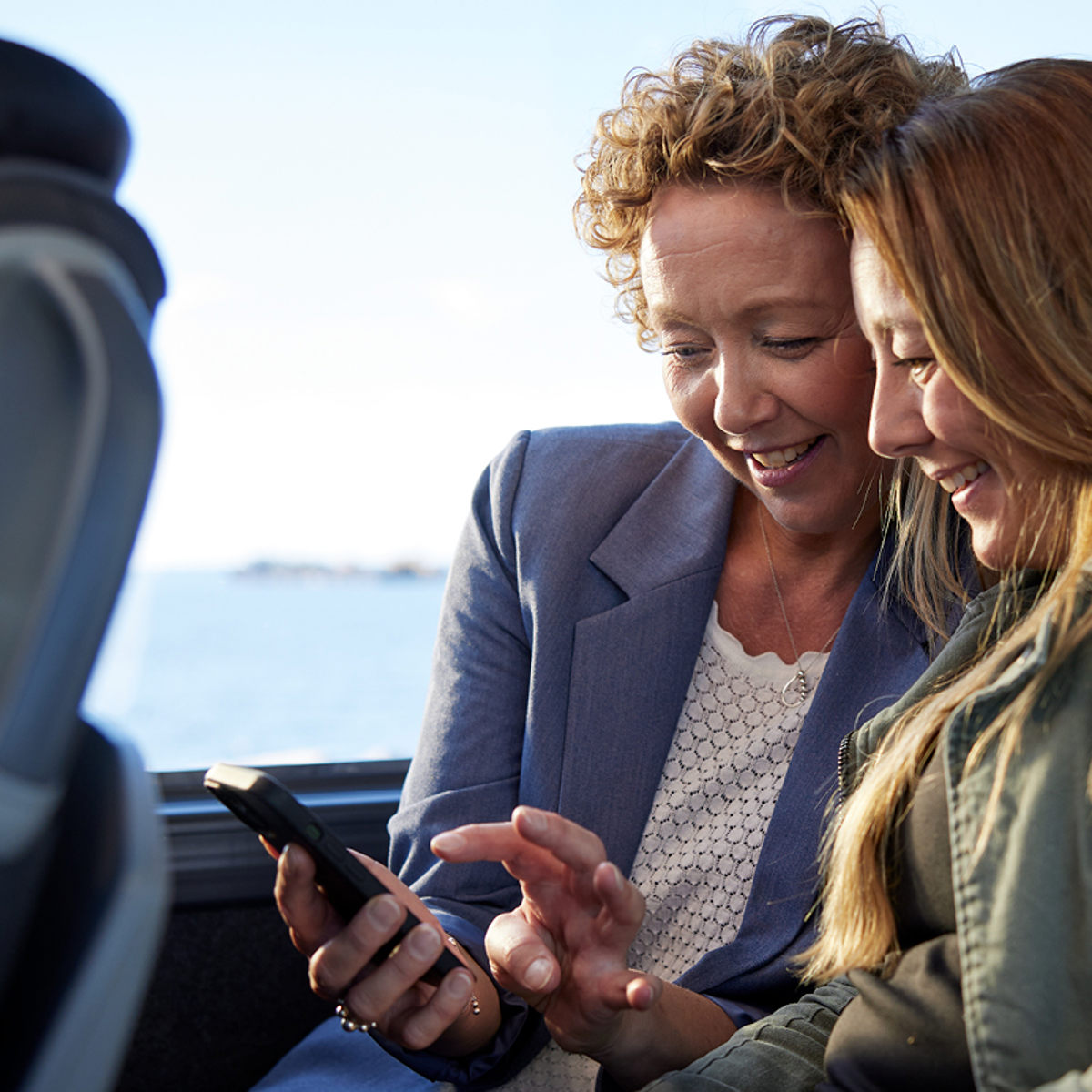 Två kvinnor ler och tittar på en telefon tillsammans på med havsutsikt genom fönstret.