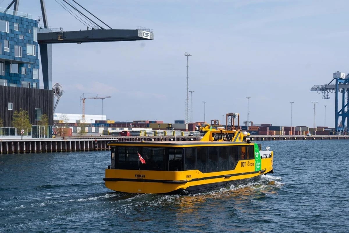 Gul hamnbåt i Köpenhamn seglar vid kajen med containerterminal i bakgrunden.