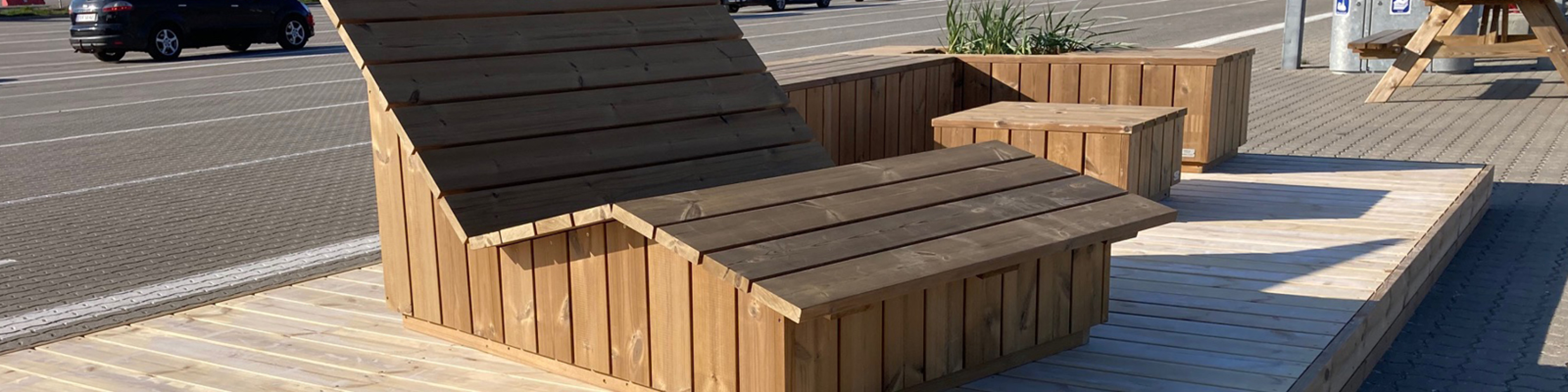 Liggestol og bord i lyst træ placeret på terrasse i et udendørsområde nær parkeringsplads.