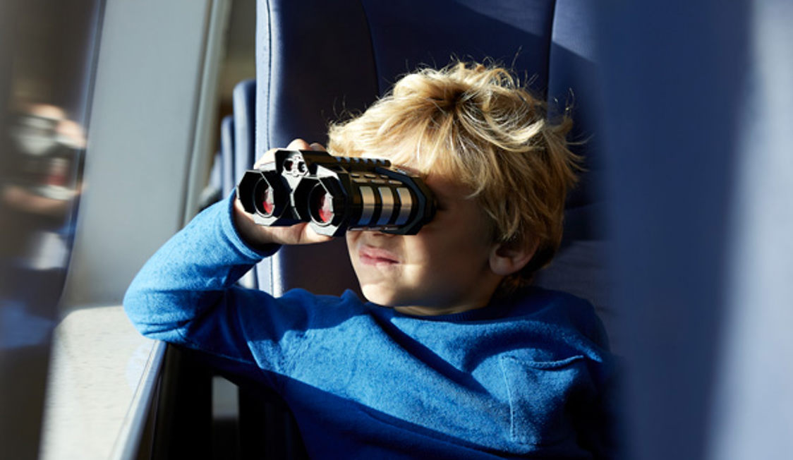 Dreng i blå trøje kigger gennem kikkert ud af et togvindue i sollys.