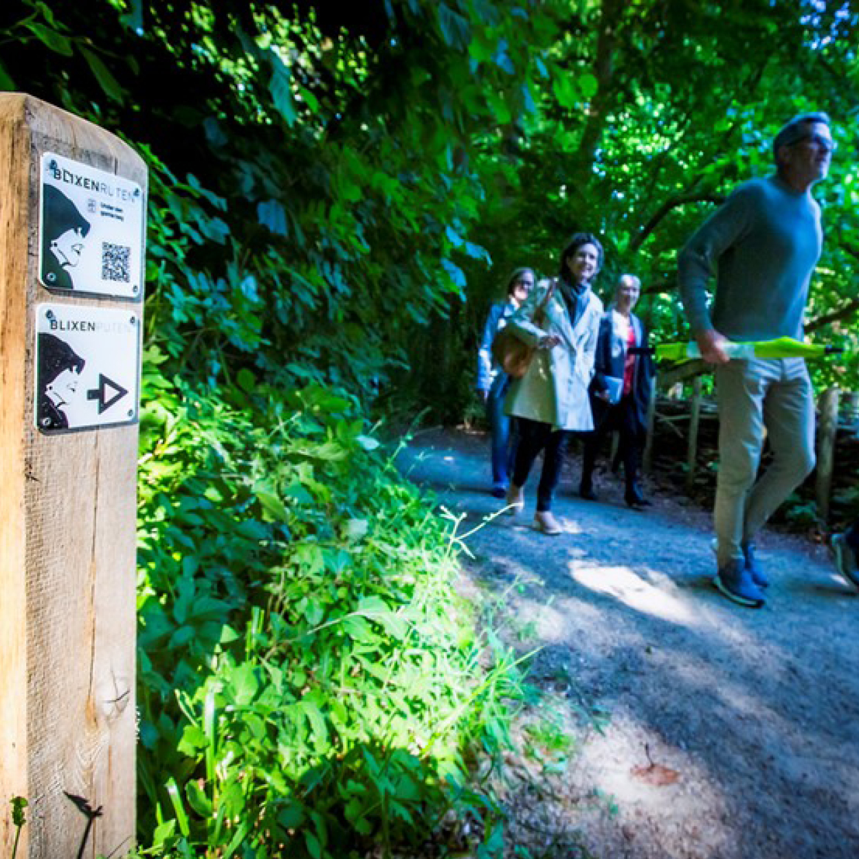 Vandrere på Blixenruten går ad skovsti, mens vejskilt med Blixenruten-logo står i forgrunden.