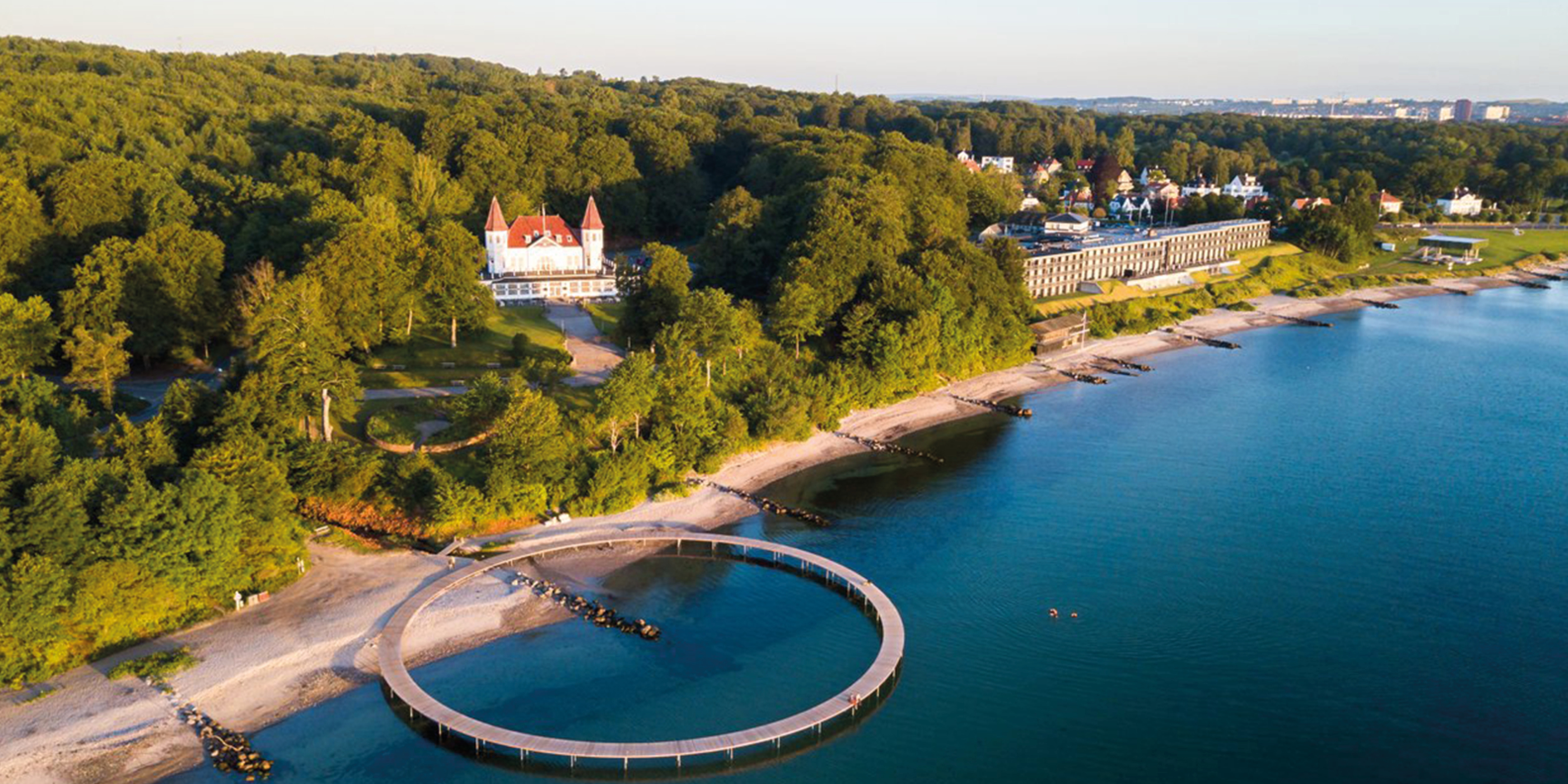 Rund badebro ved Bellevue strand ved solnedgangsvand, omgivet af frodig skov og historiske bygninger.