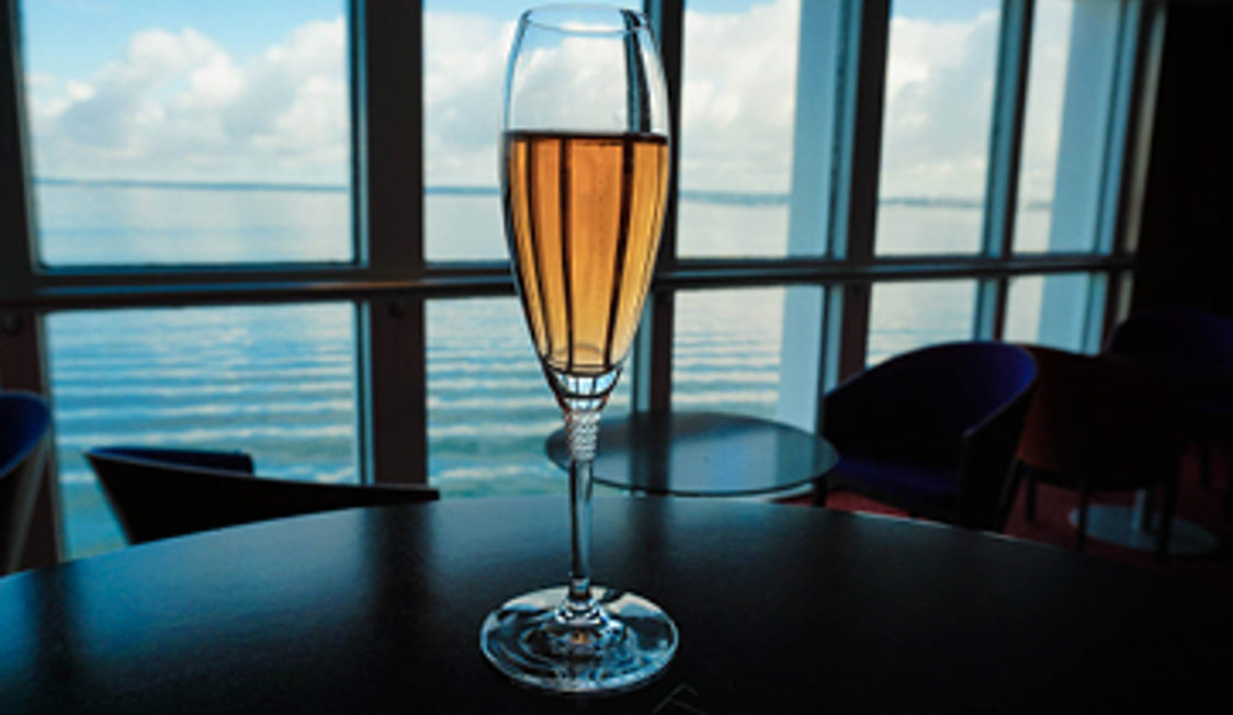 Champagneglas med gylden væske står på et rundt bord, med havudsigt gennem store vinduer i baggrunden.