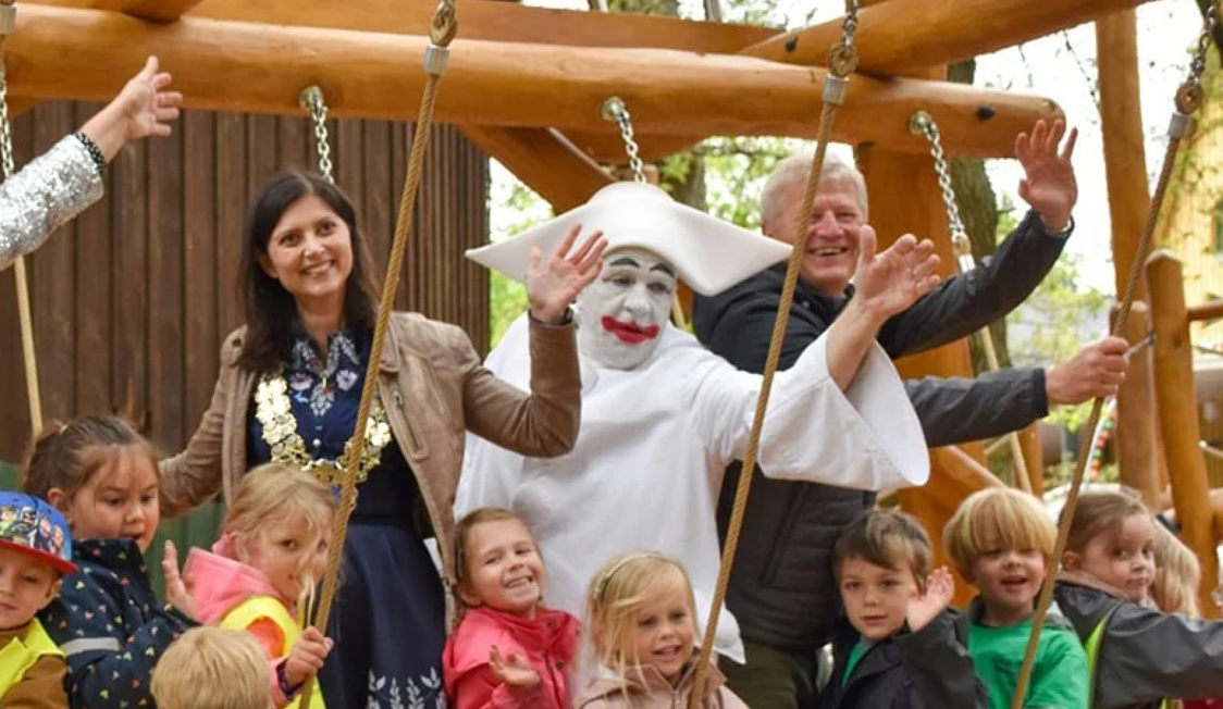 Grupp av barn och vuxna, inklusive en clown, ler framför en lekplats i skogen.