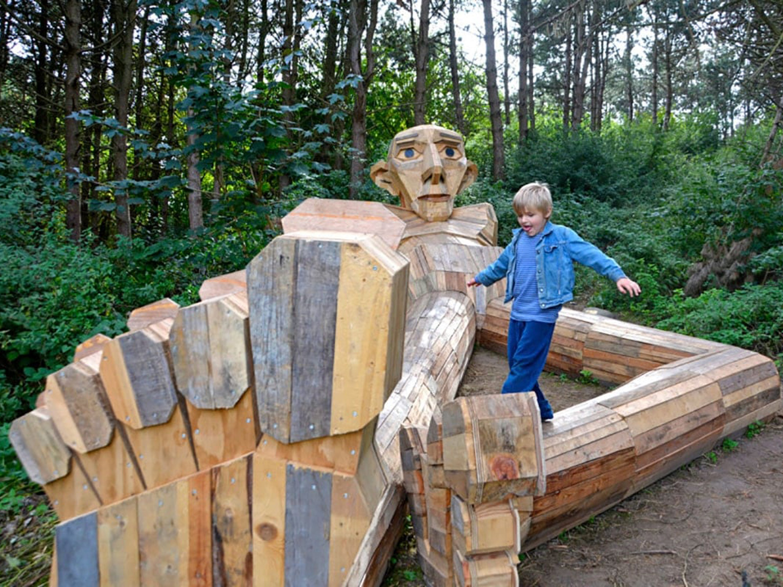 Jätteskulpturen Thomas i trä med barn som leker, omgiven av skog och grönska.