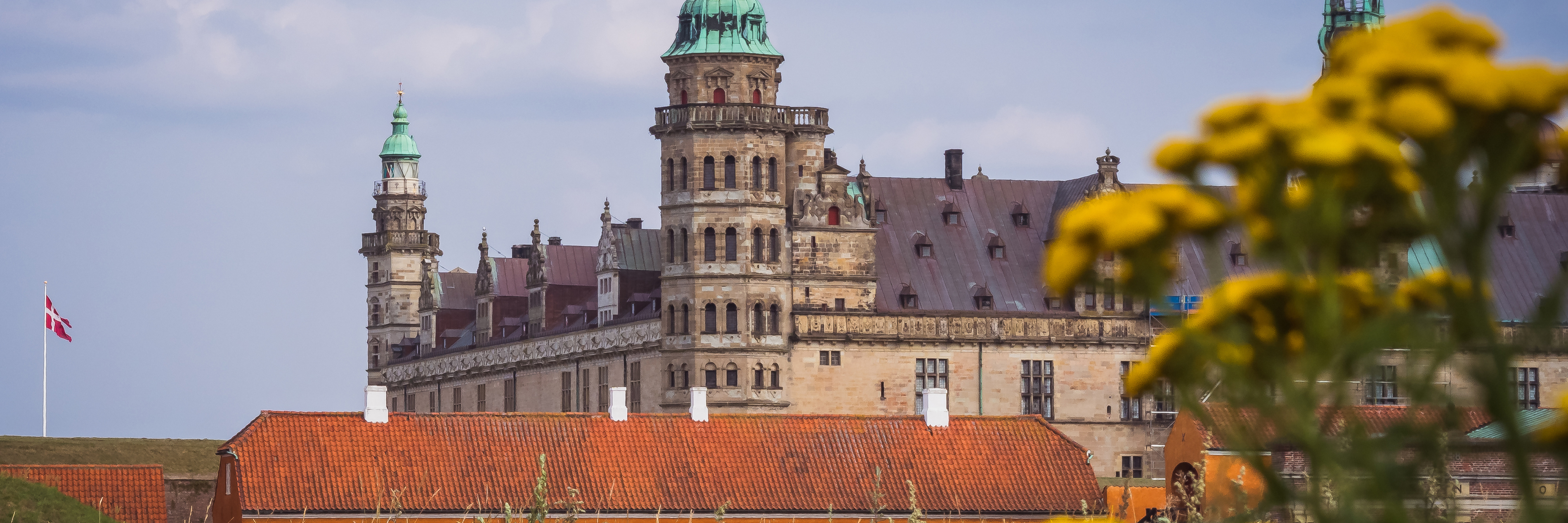 Kronborgs slott i Helsingör med danska flaggan och gula blommor i förgrunden under en klarblå himmel.