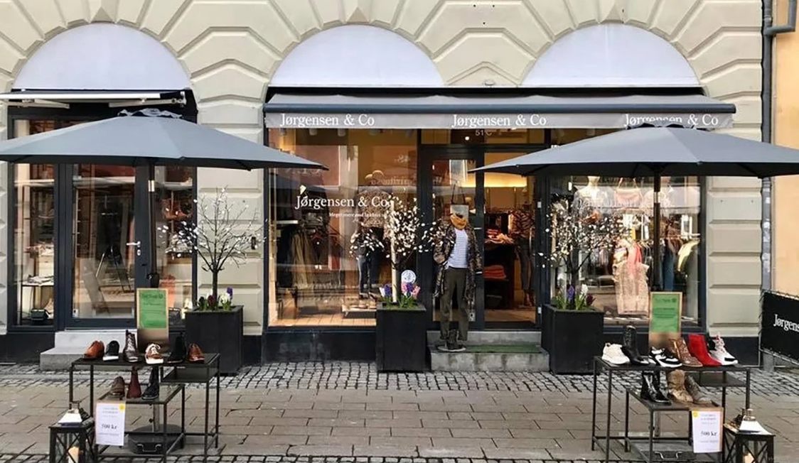 Butiksfasad på Jørgensen & Co med vårmode på dockor och skor presenterade under grå parasoller på trottoaren.